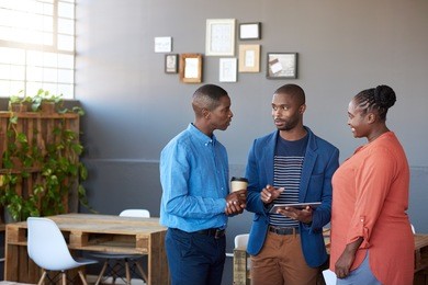 three young african office coworkers smiling and talking together over a digital tablet while standing in a large modern office