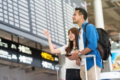 asian couple traveler holding the smart mobile phone and pointing at the flight information screen in modern an airport, travel and transportation with technology concept.