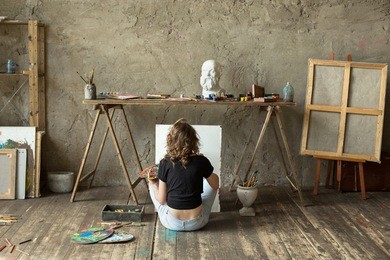 woman painter sitting on the floor in front of an empty canvas and drawing. art studio interior. horizontal background. drawing supplies, oil paints, artist brushes, canvas, frame. creative concept