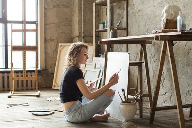 woman painter sitting on the floor in front of the canvas and drawing. artist studio interior. drawing supplies, oil paints, artist brushes, canvas, frame. workshop or art class. creative concept