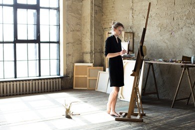 woman painter holding a brush and palette with oil paints in her hand, standing in front of an empty canvas, drawing. artist studio interior with easel and window on the background. creative concept