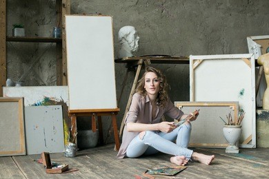 woman painter sitting on the floor among empty frames, holding paintbrush, smiling and looking at camera. artist at work. studio interior with drawing supplies on background. creative concept