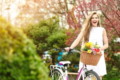 young smiling girl standing near bicycle with basket of flowers in park