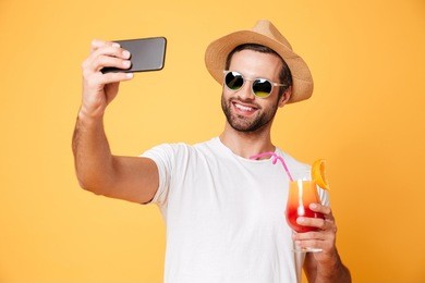 image of smiling young man standing isolated over yellow background. looking aside make selfie by mobile phone holding cocktail.