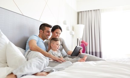 people, family and technology concept - happy mother, father and little boy with tablet pc computer in bed at home or hotel room