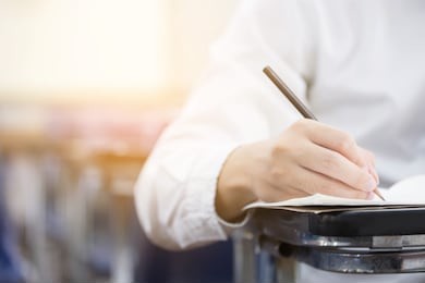 soft focus front view undergraduate holding pencil writing on paper answer sheet and sitting on lecture chair doing final exam attending in examination room or classroom.university student in uniform.