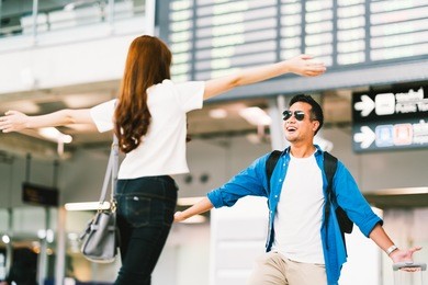 asian girl picking up her boyfriend at airport's arrival gate, welcomes back home from studying or working abroad. young couple love and hug, honeymoon, or traveling concept