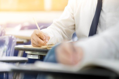 front view undergraduate student holding pencil.sitting on lecture chair doing final exam attending in examination room or study in classroom.university student in uniform.space for text.