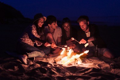 young and cheerful friends sitting on the beach and fry marshmallows near bonfire they look happy and smiling. night time