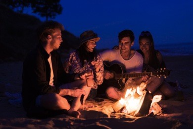 young and cheerful friends sitting on beach and fry sasuages or weenies in bonfire one man is playing guitar. music on wild beach. happy couple relationships.