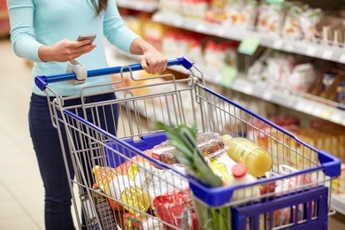 consumerism, technology and people concept - woman with smartphone and shopping cart or trolley buying food at grocery store or supermarket
