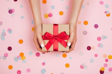 woman hands holding present box with red bow on pastel pink background with multicolored confetti. flat lay style.