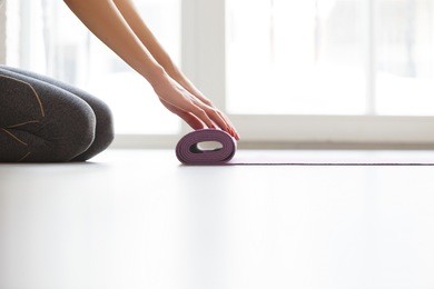 young yoga woman rolling her lilac mat after a yoga class on wooden floor near a window, close up