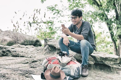 asian man happiness using smartphone sitting alone on rock in forest listening music earphones. having rest outdoor. nature and modern technologies and lifestyle communications concept