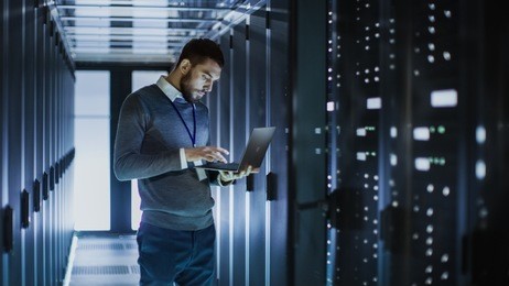 it technician works on laptop next to a server cabinet in big data center. he runs diagnostics and maintenance, sets system up.