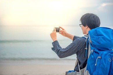 young asian backpacker man taking photos of beach and sea by smartphone, summer holiday vacation and travel tropical island concepts