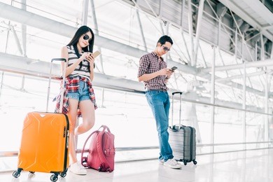 young asian man and woman using smartphone checking flight or online check-in at airport together, with luggage. air travel, summer holiday, or mobile phone application technology concept