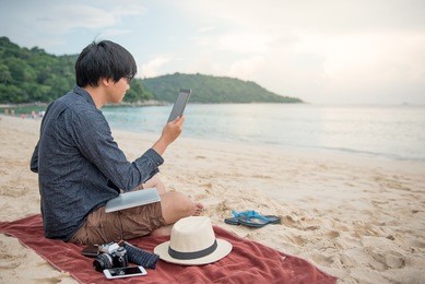 young asian man working using tablet on tropical beach, digital nomad lifestyle or freelance occupation concepts