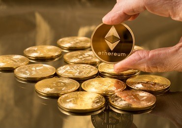 hand holding a single ether or ethereum coin over bitcoins on gold background to illustrate blockchain and cyber currency