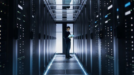 male it engineer works on a laptop at the end of a corridor in a big data center. rows of rack servers are seen.
