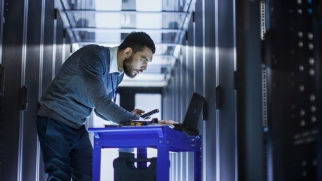 it engineer with tool cart working on a laptop computer, he holds a hard drive. he stands at a corridor of a large data center full of rack servers.