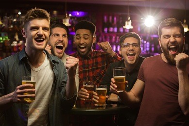 four happy men holding beer mugs and gesturing while watching tv in bar or pub