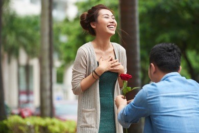 portrait of man proposing to smiling girlfriend in street, standing on one knee and holding red rose