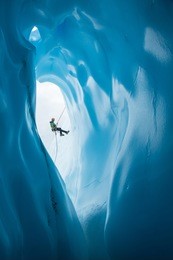 an ice climber in a green jacket and orange helmet rappels past a large rounded entrance to an ice cave on the matanuska glacier in alaska.
