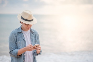young asian man with jean shirt and hat holding smartphone and checking social media application on tropical beach at sunset, background for summer holiday vacation or global communication concepts