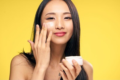 asian, woman on yellow background applies cosmetic face cream.