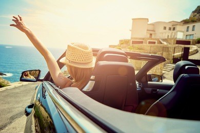 back view of blonde in straw hat driving cabriolet with blue sea and houses on background.