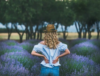 young blond woman traveller wearing straw hat, denim shorts and blue off-shoulder blouse standing in lavender field surrounded with lavender flowers in isparta region of turkey