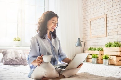 happy casual beautiful woman working on a laptop sitting on the bed in the house.