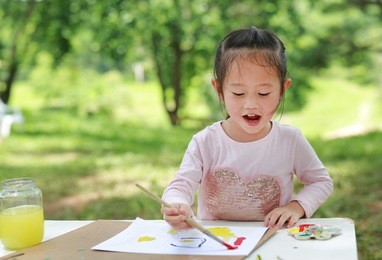 cute asian child girl sitting at a table in a summer garden painting with paintbrush