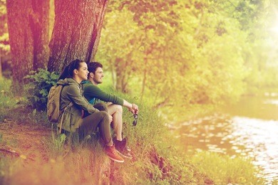 travel, hiking, backpacking, tourism and people concept - smiling couple with backpacks resting on river bank in nature on river bank