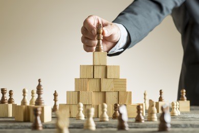 businessman placing a chess piece on a pyramid of wooden building blocks in a concept of success and achievement in a close up view of his arm.