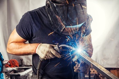 a strong man is a welder in a black t-shirt, in a welding mask and welders leathers, a metal product is welded with a welding machine in the garage, blue sparks fly to the sides