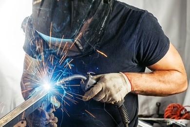 a strong man welder in a black t-shirt, in a welding mask and welders leathers weld metal welding machine in the workshop, in the sides fly the blue sparks