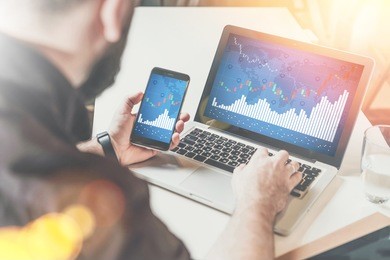rear view. young bearded businessman sitting at desk and using laptop while holding smartphone with graphs and charts, diagrams on screen. online marketing, education, startup. hipster man working