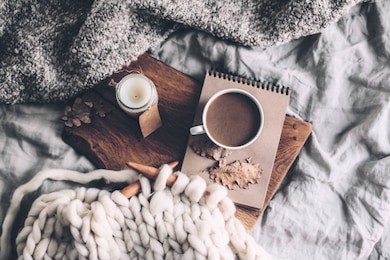 cup of coffee and candle on rustic wooden serving tray in the cozy bed with blanket. knitting warm woolen sweater in the autumn weekend, top view.
