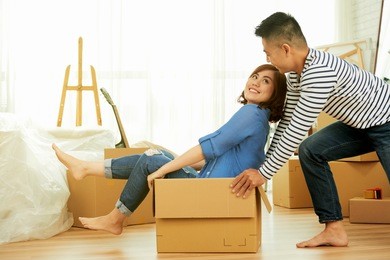 profile view of lovely asian couple having fun while riding in cardboard box at new apartment, interior design items and moving boxes on background