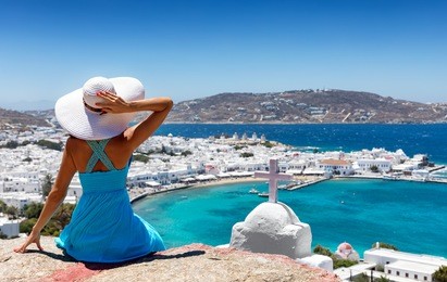elegant woman with blue dress and white hat enjoys the view over the town of mykonos, greece