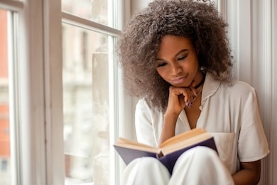 pretty afro-american girl with reading a book sitting on the windowsill.