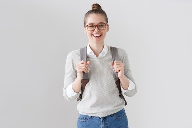 portrait of energetic teenager girl isolated on gray background pulling forward straps of gray hipster backpack, laughing happily as if anticipating friendly meeting or great leisure time in open air.