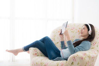 smiling happy girl wearing earphones looking at mobile digital tablet and lying down on sofa chair enjoying holiday vacation.