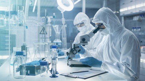 in a secure high level laboratory scientists in a coverall conducting a research. chemist adjusts samples in a  petri dish with pincers and then examines them under microscope.