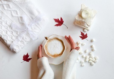 autumn composition. hot chocolate, knitted blanket, lit candle, autumn leaves. flat lay, top view, white background. hands holding cup of warm drink