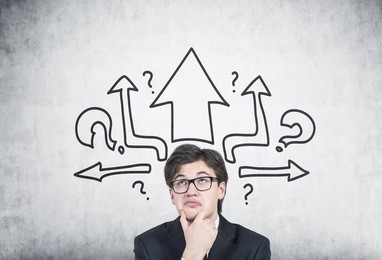 close up portrait of a pensive young businessman wearing glasses and a black suit and analysing while standing near a concrete wall with arrows and question marks on it.