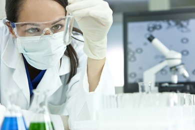 a female medical or scientific researcher or woman doctor looking at a test tube of clear solution in a laboratory with her microscope beside her.