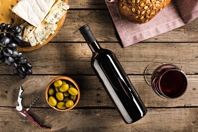 top view of bottle of red wine, corkscrew, wineglass, green olives, different cheese and bread on napkin on wooden tabletop